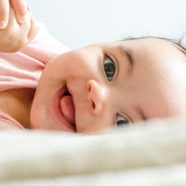 A smiling baby lies on a soft surface, wearing a pink shirt. The background is a light blur, adding a serene and joyful mood.