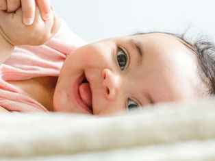 A smiling baby lies on a soft surface, wearing a pink shirt. The background is a light blur, adding a serene and joyful mood.