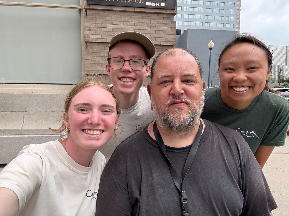 Four people smiling in a city setting. Three wear "Christ in the City" shirts. There's a light pole and buildings in the background.