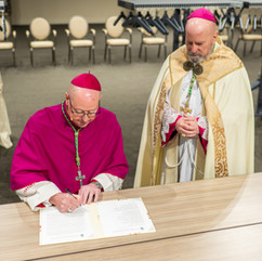 The bishop signs a document at a table while another clergyman in cream-colored vestments stands beside him with hands folded in prayer.