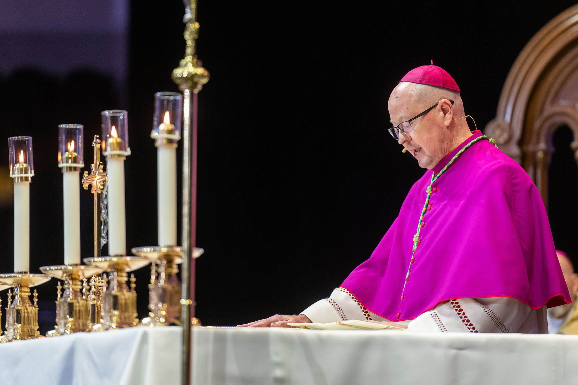 The bishop reads from a liturgical book at an altar surrounded by lit candles, wearing magenta vestments.