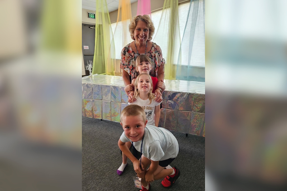 Mujer sonriente con tres niños en sala decorada con telas de colores y mesa brillante; ambiente alegre.