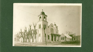 Sepia photo of a white church with bell towers and crosses on a quiet street, framed by leafless trees on a green background.