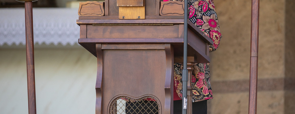 Elderly woman in floral jacket speaks at wooden podium with candles and microphone in a solemn setting.