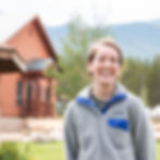 Smiling person in a gray fleece stands outdoors against a rustic cabin and mountain backdrop. Bright and cheerful mood.