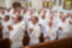 Clergy in white robes stand in a church, hands clasped in prayer. The setting is formal, with gold decor on the walls. Mood is reverent.