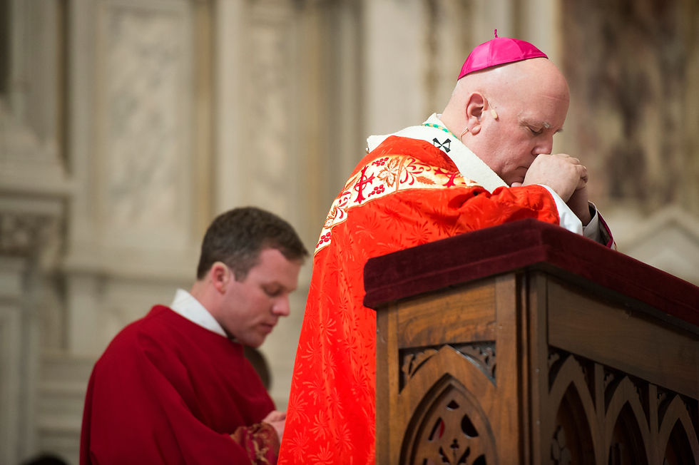 Two clerics in red robes pray in a cathedral. One has a pink cap, both appear solemn. Ornate wooden pulpit in foreground.