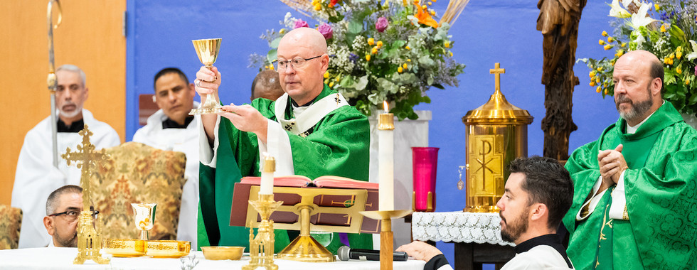 Bishop in green vestments lifts a chalice at a Catholic altar with candles, flowers, crucifix, and seated clergy.