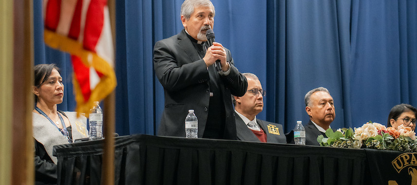 A man speaks at a podium with a microphone, flanked by seated people. An American flag is visible. Blue curtains in the background.
