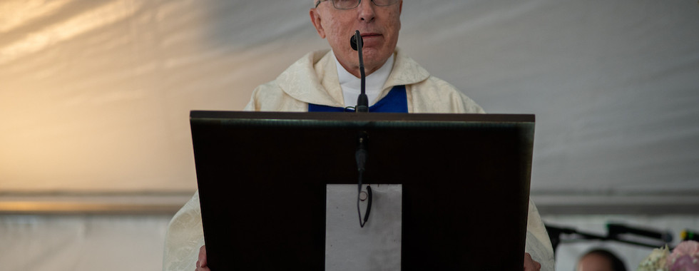 A priest in white vestments speaks from a lectern during a church gathering inside a tent.