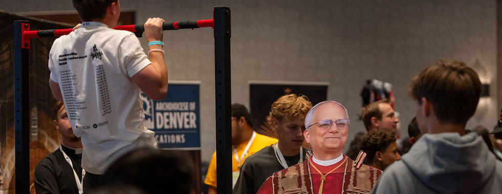 Teen does a pull-up on a red bar in a crowded room; a cardboard bishop cutout stands by a Denver sign.