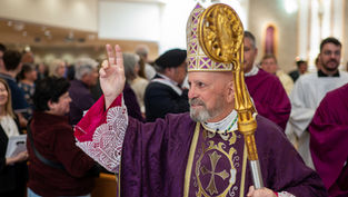 Bishop in ornate purple robe and mitre holds up two fingers in blessing. He carries a golden staff. People gather in a church setting.