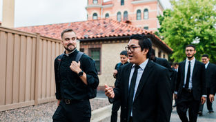Group of men in suits and clerical attire walking and chatting outside, with brick building and greenery in the background. Casual mood.