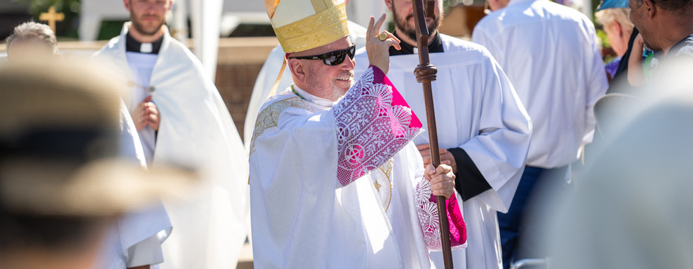 Bishop in white vestments and gold mitre smiles, holding a staff and blessing outdoors amid other clergy and onlookers.