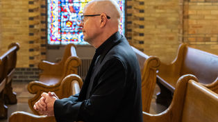 A person in a black clerical outfit prays in a church pew, with colorful stained glass in the background. The setting is solemn and introspective.