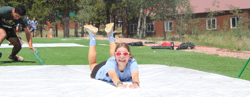 Smiling woman slides belly-first on a wet slip-and-slide as water sprays, with a man at left, on a grassy field by trees and cabins.