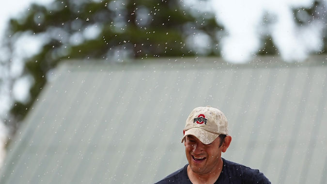 Man in a cap and race number 20 crosses a finish line, smiling in the rain. Background shows trees and a green-roofed building.