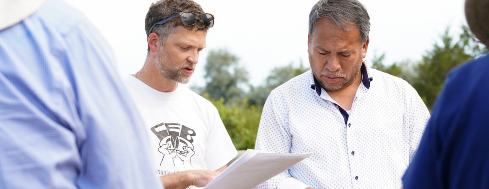 Two men outdoors study a paper, one holding rosary beads, with blurred people around them under a bright sky.