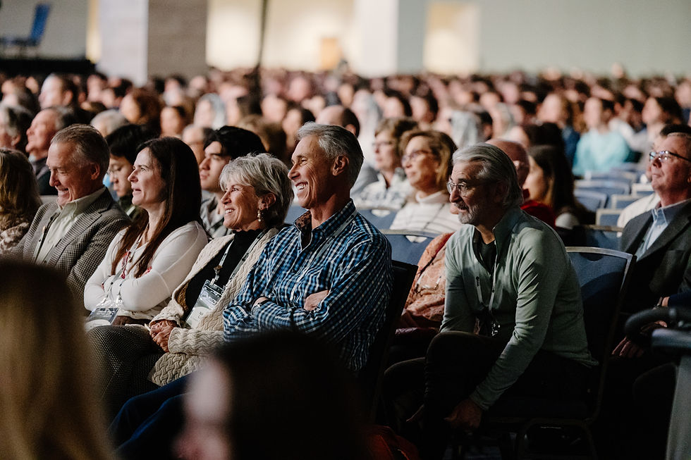 Audience seated in a large hall, smiling and attentive. Rows of people create a sense of community and interest. Neutral background.