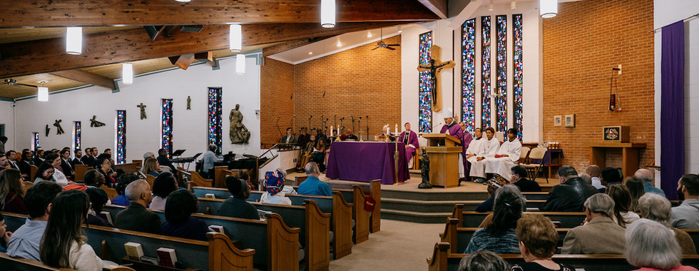 Congregation in a church service with a bishop in purple robes at the altar, surrounded by clergy. Stained glass windows and wooden beams.