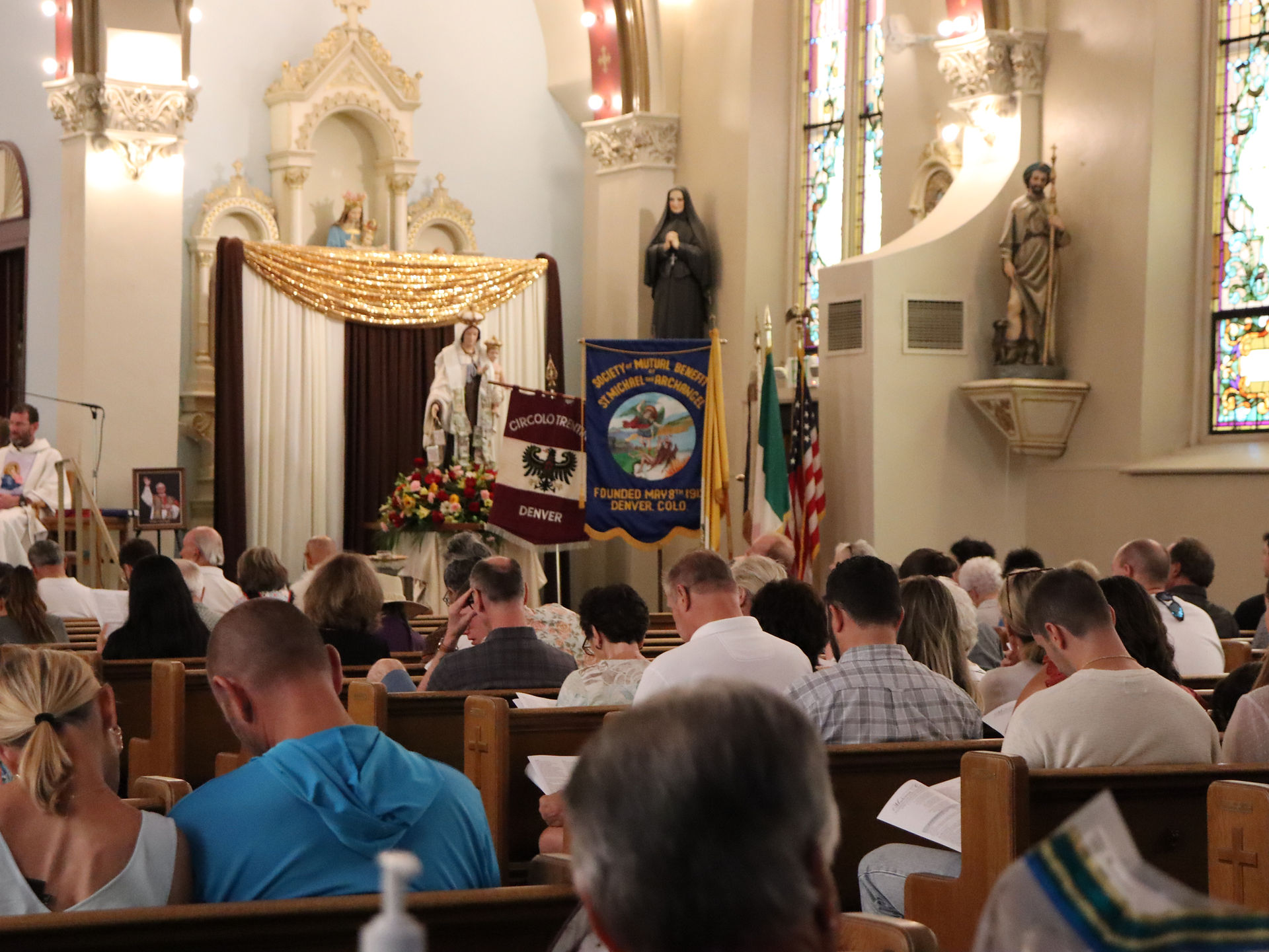Congregation seated in a decorated Catholic church during Mass, with banners reading Denver and St. Michael, softly lit and solemn