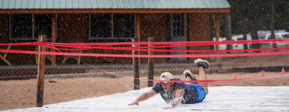 Man in blue slides on wet tarp under red ribbons. Background: wooden cabin, green roof, trees. Light rain, playful mood.