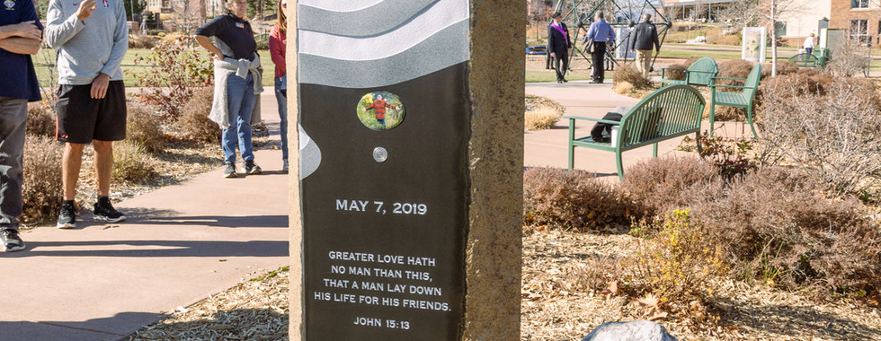 A stone memorial with an engraved American flag and scripture is in a park. People stand observing. Blue sky, brown path, and buildings behind.