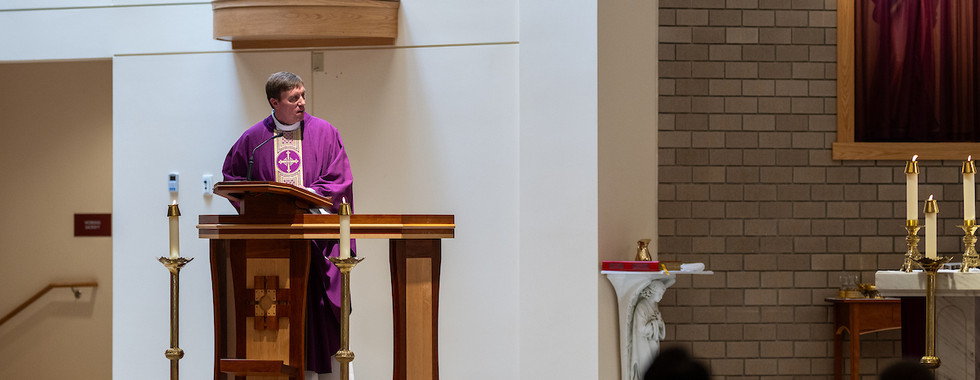 A priest in a purple robe speaks at a wooden lectern in a church. Candles and altar visible; audience blurred in foreground. Calm atmosphere.