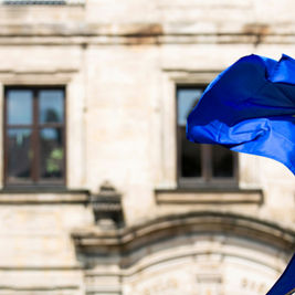 European Union flag with yellow stars waving in the wind in front of a historic stone building with arched windows.