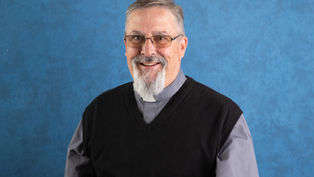 Hombre con barba y gafas sonriendo, vestido con camisa gris y chaleco negro, sobre fondo azul. El ambiente es amigable.