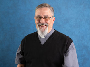 Hombre con barba y gafas sonriendo, vestido con camisa gris y chaleco negro, sobre fondo azul. El ambiente es amigable.