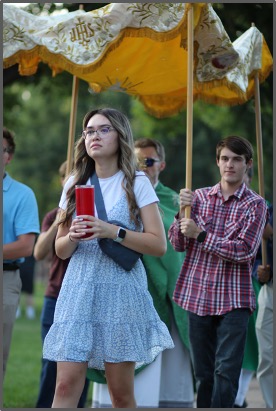 Young woman in a blue dress holds a red candle, walking under a yellow canopy. Two men follow, holding the canopy. Green park setting.