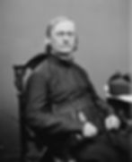 Black-and-white portrait of a solemn seated clergyman in dark robes, looking at the camera against a plain backdrop.
