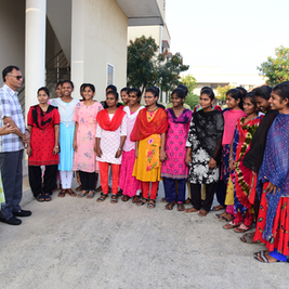 Smiling women and a man stand in a semicircle outside a building, wearing colorful salwar kameez and chatting.