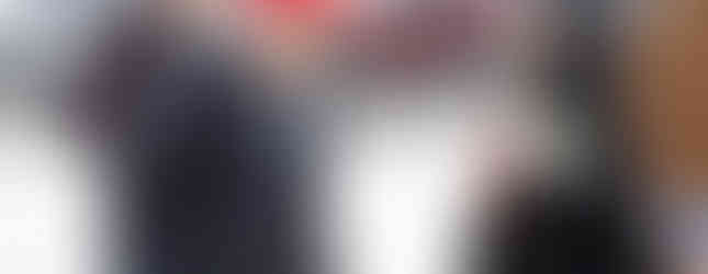 Man in a red cap talks with a smiling nun holding a book and bottle. Background shows a fence, cars, and a woman in brown coat.