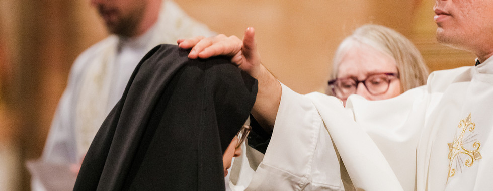 Priest in white robe touches a nun's black veil during a ceremony, with another person in the background. Warm, solemn atmosphere.