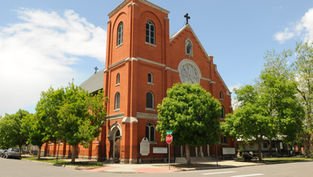 Red brick church with arched windows, cross on top, and trees in front. A stop sign is visible; it's a sunny day with a few clouds.