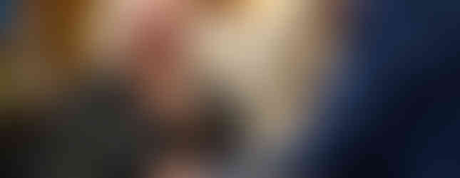 Elderly man in black clerical attire, gesturing while talking to a person in blue suit. They're seated in a cozy room with blurred bookshelves.