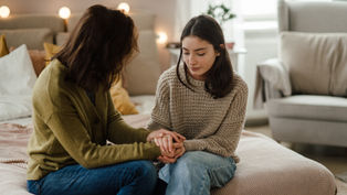 Two women sit on a bed holding hands in a cozy room with soft lighting. One wears a green cardigan, the other a beige sweater, both appear thoughtful.