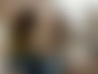 Two women sit on a bed holding hands in a cozy room with soft lighting. One wears a green cardigan, the other a beige sweater, both appear thoughtful.