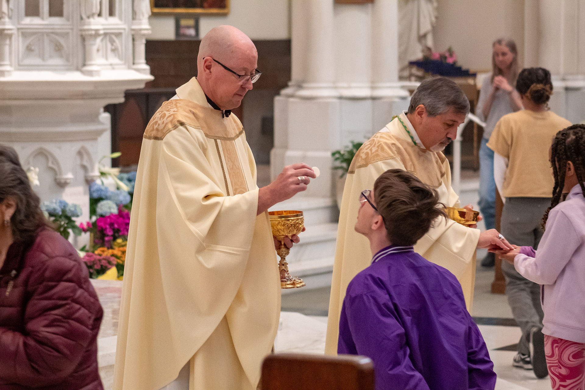 A bishop gives Communion to a kneeling young man as another minister stands nearby.