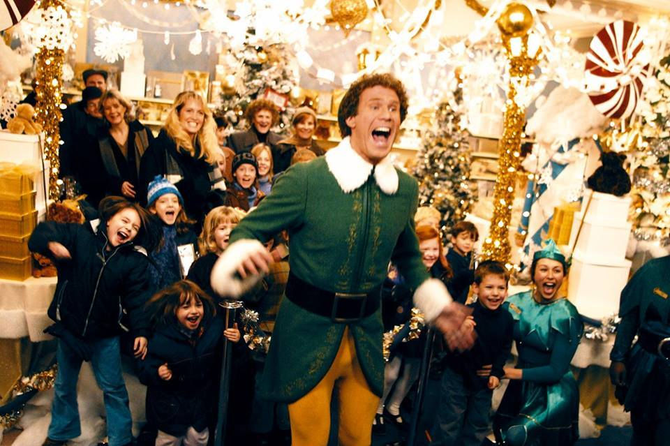 Elf-costumed man joyfully leads excited crowd in festive, decorated store with Christmas trees, lights, and gifts; cheerful atmosphere.