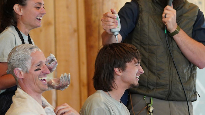 Four people in a wooden room, laughing. One holds a mic, others with hair bleach on heads. Casual, joyful atmosphere.