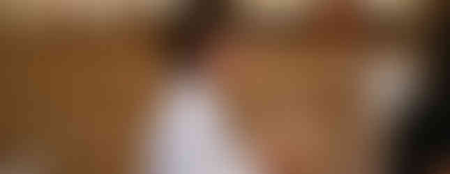 A woman in a white coat holds a glass vessel in a room with wooden paneling. Subtle lighting and blurred background suggest focus.