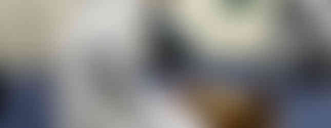 Person in white attire kneeling on blue carpet, meditating near a wooden stand with books. A seated person in blue is in the background.