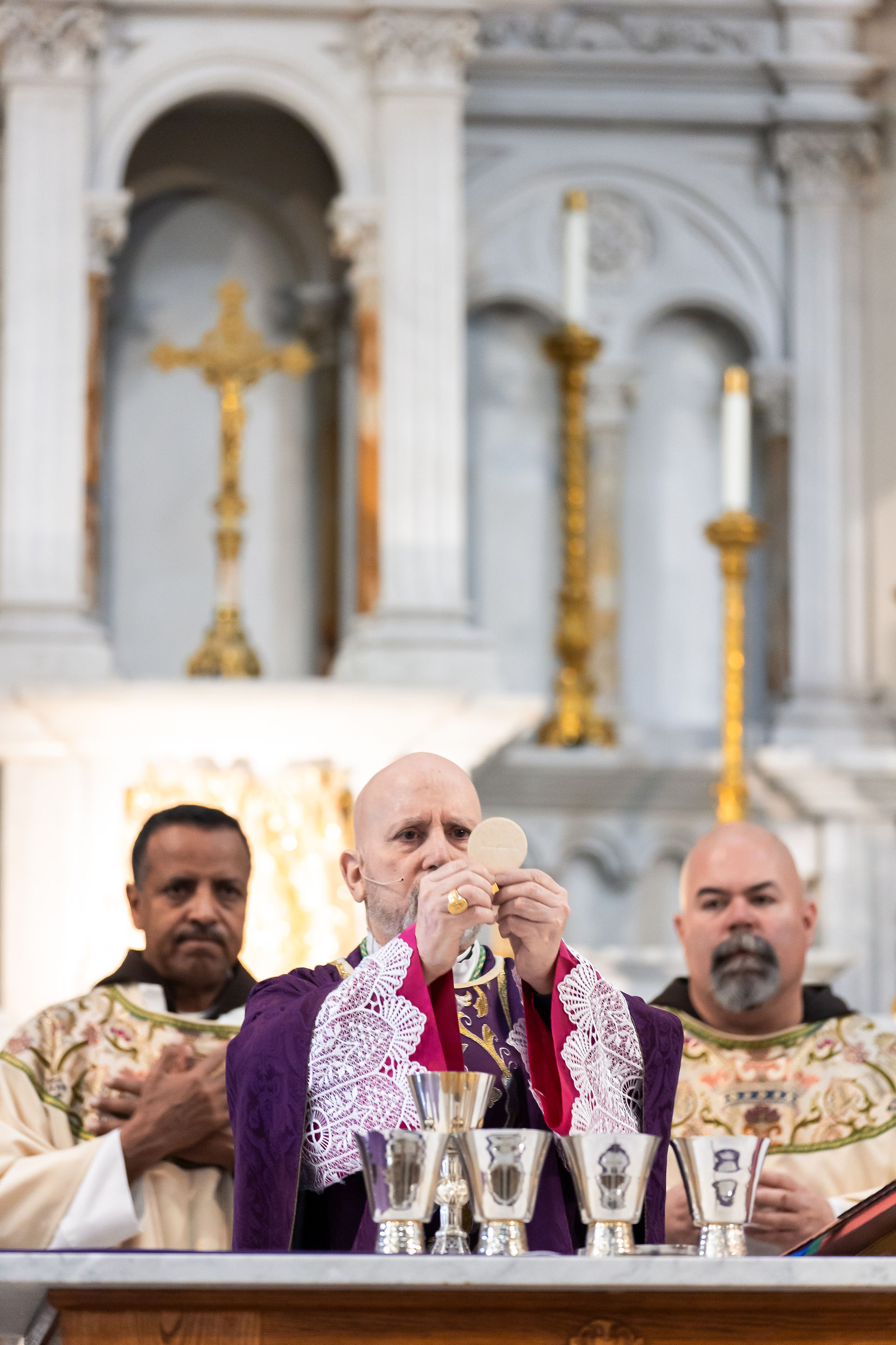 Bishop in purple robe holds up Eucharist during mass, flanked by two men in vestments. Ornate altar with candles and gold crucifix behind.