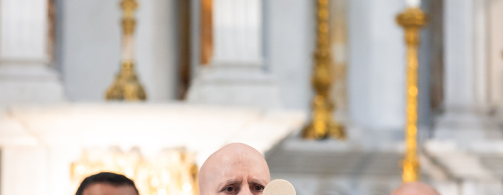 Bishop in purple robe holds up Eucharist during mass, flanked by two men in vestments. Ornate altar with candles and gold crucifix behind.