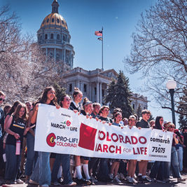 Personas sostienen pancartas "Colorado March for Life" frente a un edificio con cúpula dorada. Ambiente soleado y de protesta pacífica.