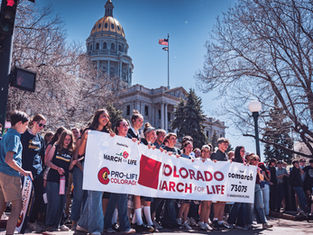 Personas sostienen pancartas "Colorado March for Life" frente a un edificio con cúpula dorada. Ambiente soleado y de protesta pacífica.
