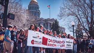 Personas sostienen pancartas "Colorado March for Life" frente a un edificio con cúpula dorada. Ambiente soleado y de protesta pacífica.